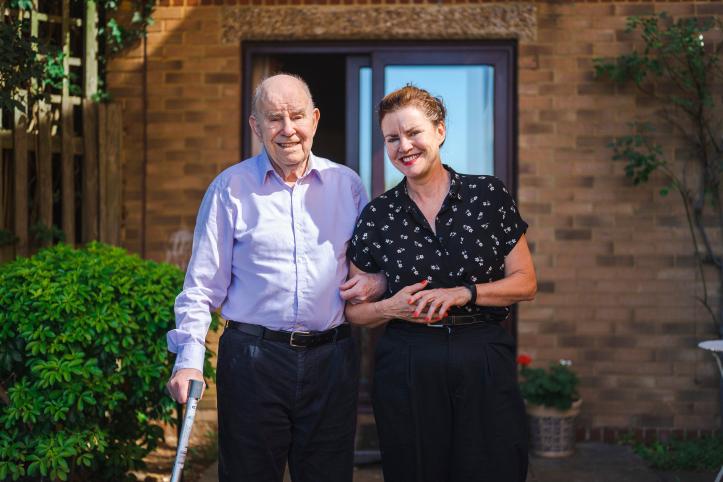 A father and daughter walking outside a house, arm in arm. The father is using a walking stick.